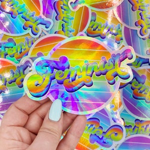 May include: A holographic sticker with the word "Feminist" in a retro font on a rainbow striped background.