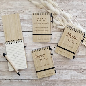 Peut inclure: Plusieurs blocs-notes en bois avec reliure spirale et stylos attachés. Les blocs-notes comportent des inscriptions gravées en français, dont "Merci" et des messages personnalisés. Un bloc-notes est ouvert, révélant du papier ligné et un cœur dessiné.