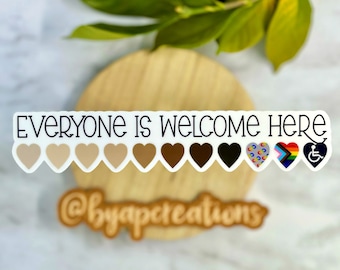 Puede incluir: Una pegatina blanca con el texto "Everyone is welcome here" y una fila de corazones en diferentes tonos de piel, seguidos de corazones con banderas de orgullo arcoíris, de género y de discapacidad. La pegatina también incluye el texto "@byapcreations".