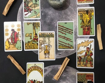 Puede incluir: Tirada de lectura de cartas de tarot con diez cartas dispuestas sobre un fondo oscuro. Las cartas son de la baraja de tarot Rider-Waite e incluyen el Caballero de Espadas, el As de Copas, la Reina de Bastos, el Paje de Bastos, el Diez de Bastos y la carta del Mundo.
