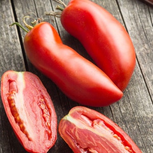 May include: Two whole elongated red tomatoes with green stems and two halved tomatoes showing internal seeds and pulp, arranged alongside a kitchen knife with a wooden handle on a weathered gray wooden surface.