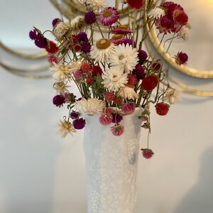 May include: A white milk glass vase with a textured design holds a bouquet of dried flowers. The flowers are white, pink, and purple. The vase is on a white surface.