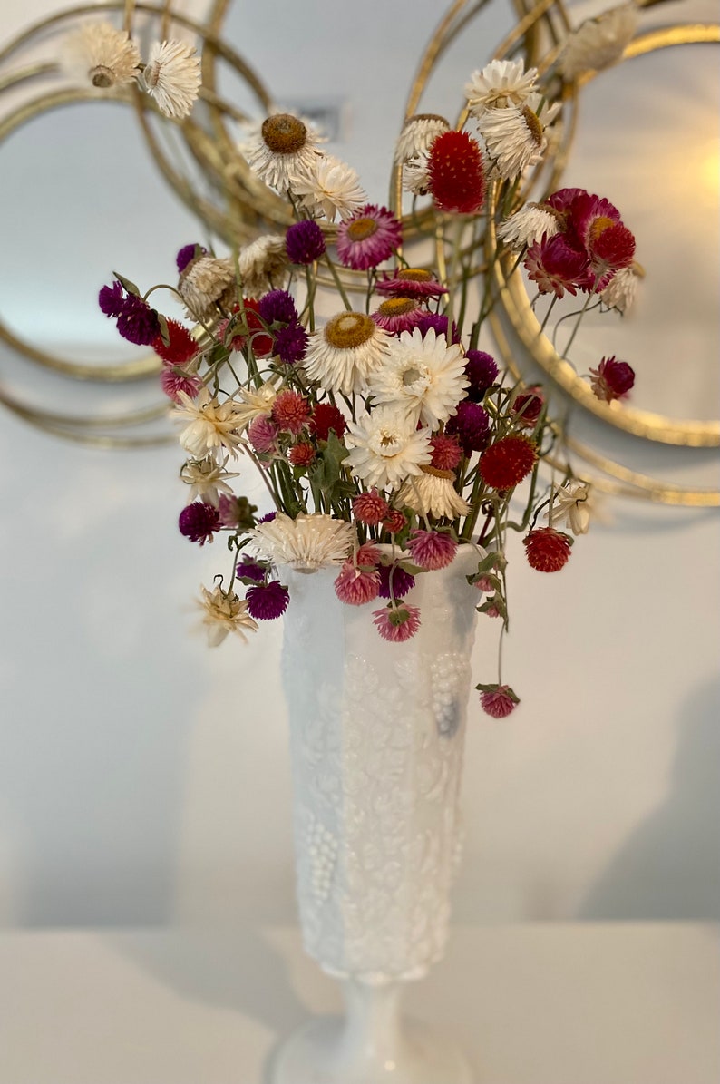May include: A white milk glass vase with a textured design holds a bouquet of dried flowers. The flowers are white, pink, and purple. The vase is on a white surface.