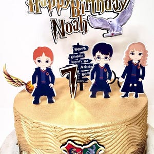 May include: A gold and brown cake decorated with a Harry Potter theme. The cake has a golden Hogwarts crest, a golden number 7, and figurines of Harry Potter, Ron Weasley, and Hermione Granger. The cake also has a sign that says "Hogwarts, Gringotts, Hagrid's Hut, Azkaban". The cake says "Happy Birthday Noah" on top.