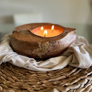 May include: A three-wick candle burning in a natural coconut shell holder. The candle is surrounded by a white cloth and a woven basket.
