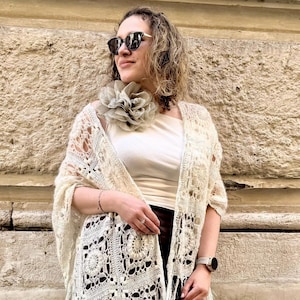 May include: A cream-coloured, open-weave shawl with a fringed hem. The shawl features a floral crochet pattern and is worn over a white top and brown skirt. A large, grey flower pin adorns the shoulder.