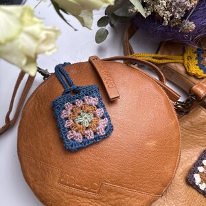 May include: A round tan leather bag with a small square crochet granny square charm attached. The charm features a floral pattern in shades of blue, pink, green, and brown. The bag has the brand name muud embossed on the front leather surface.