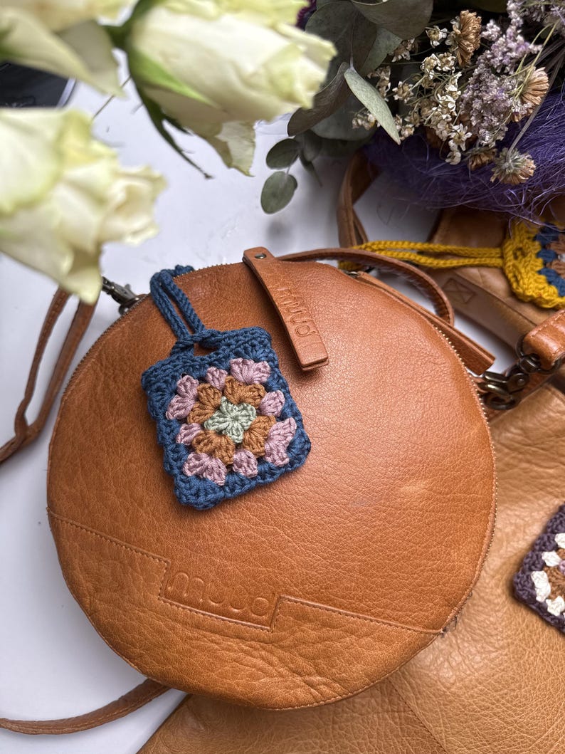 May include: A round tan leather bag with a small square crochet granny square charm attached. The charm features a floral pattern in shades of blue, pink, green, and brown. The bag has the brand name muud embossed on the front leather surface.