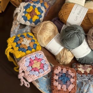 May include: A collection of multi-colored crochet granny squares featuring various yarn color combinations, including yellow, blue, pink, and brown, arranged alongside several skeins of yarn with labels reading Tencel and Soft Bamboo.