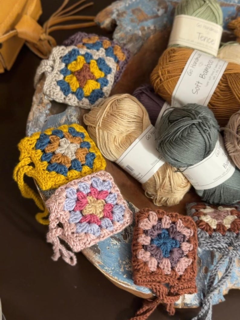 May include: A collection of multi-colored crochet granny squares featuring various yarn color combinations, including yellow, blue, pink, and brown, arranged alongside several skeins of yarn with labels reading Tencel and Soft Bamboo.