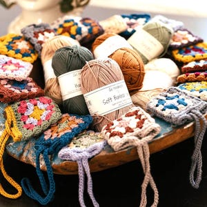 May include: A collection of small, multi-colored crocheted granny square pouches with attached loops, arranged in a round wooden bowl alongside several skeins of yarn labeled Go Handmade Soft Bamboo, featuring various earth and bright tones.