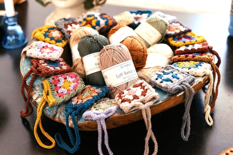 May include: A collection of small, multi-colored crocheted granny square pouches with attached loops, arranged in a round wooden bowl alongside several skeins of yarn labeled Go Handmade Soft Bamboo, featuring various earth and bright tones.