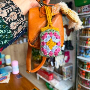 May include: A small, rectangular crochet pouch featuring a colorful granny square pattern in yellow, light blue, and pink, bordered by sage green yarn. The pouch is attached to a tan leather handbag by a mustard yellow crocheted strap.