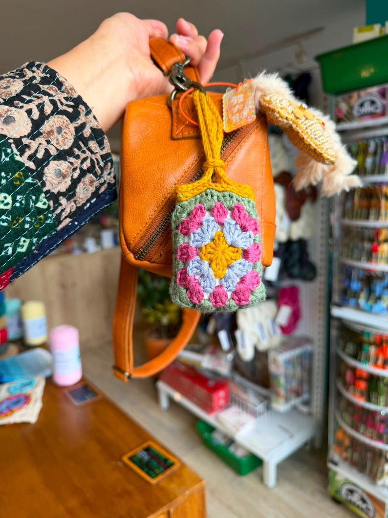 May include: A small, rectangular crochet pouch featuring a colorful granny square pattern in yellow, light blue, and pink, bordered by sage green yarn. The pouch is attached to a tan leather handbag by a mustard yellow crocheted strap.