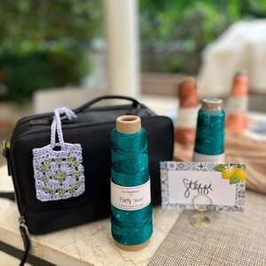 May include: A black rectangular zippered bag, a small lavender and sage green crocheted square with a handle, a spool of teal yarn labeled Go Handmade Party Deluxe, and a white rectangular place card with the name Steffi and lemon graphics.