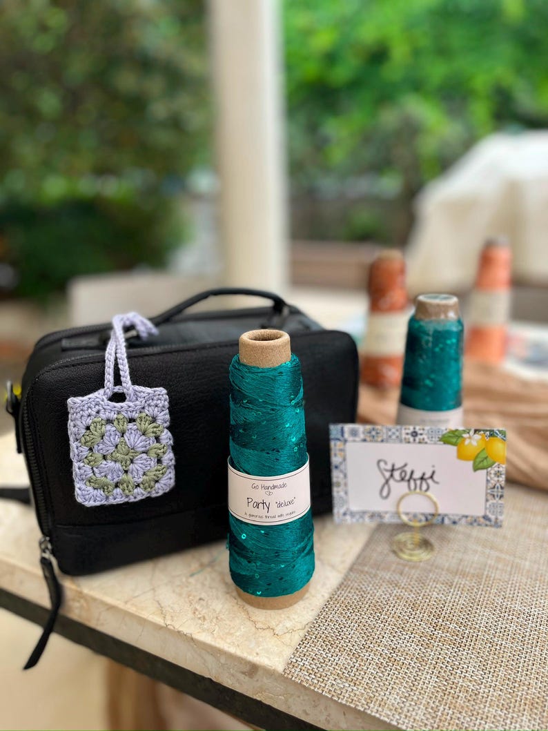 May include: A black rectangular zippered bag, a small lavender and sage green crocheted square with a handle, a spool of teal yarn labeled Go Handmade Party Deluxe, and a white rectangular place card with the name Steffi and lemon graphics.