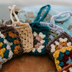 May include: A collection of small, square crochet pouches featuring various granny square patterns in colors including mustard yellow, teal, cream, brown, and dusty rose. Each pouch has a matching crocheted loop handle attached to the top edge.