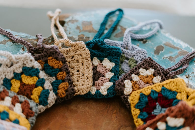 May include: A collection of small, square crochet pouches featuring various granny square patterns in colors including mustard yellow, teal, cream, brown, and dusty rose. Each pouch has a matching crocheted loop handle attached to the top edge.