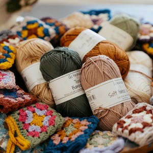May include: A collection of yarn skeins in shades of dark green, dusty pink, tan, and brown, each wrapped in a white paper label reading Go Handmade Tender Bamboo or Soft Bamboo, interspersed with small, colorful crocheted granny square motifs.