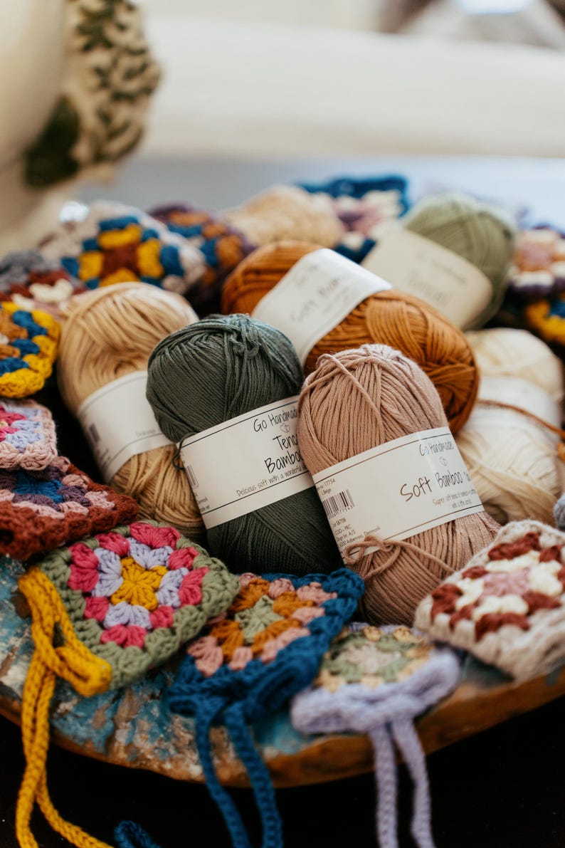 May include: A collection of yarn skeins in shades of dark green, dusty pink, tan, and brown, each wrapped in a white paper label reading Go Handmade Tender Bamboo or Soft Bamboo, interspersed with small, colorful crocheted granny square motifs.