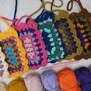 Könnte beinhalten: Eine Sammlung rechteckiger Häkeltaschen mit mehrfarbigen Granny-Square-Mustern und Trageriemen, angeordnet neben mehreren Garnknäueln in verschiedenen Farben und einer einzelnen rosa Häkelnadel auf einer weißen Oberfläche.