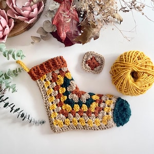 Könnte beinhalten: Ein handgehäkelter Strumpf in Gelb-, Orange- und Türkistönen mit brauner Manschette. Ein kleines quadratisches Häkelstück und ein Knäuel gelber Wolle liegen neben dem Strumpf. Der Hintergrund umfasst getrocknete Blätter und Grünzeug.