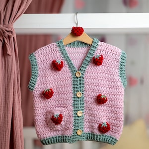 Peut inclure: Un gilet rose en crochet avec des bordures vertes et des boutons en bois. Le gilet est orné d'applications de fraises rouges. Il est suspendu à un cintre en bois avec un cœur rouge. Le gilet est sans manches.