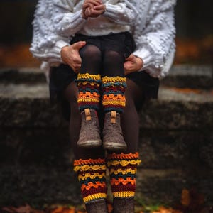 May include: A child and adult wearing matching brown boots with colorful, crocheted leg warmers. The leg warmers feature a pattern of yellow, orange, and teal. The child is wearing a white sweater and a brown beanie.