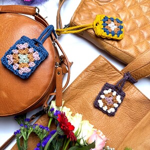 May include: Three square crochet granny square keychains with wrist straps, each featuring a unique color combination of blue, yellow, or purple yarn, attached to three distinct tan leather handbags arranged on a white surface with flowers.