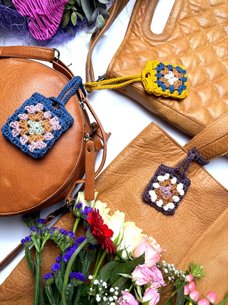 May include: Three square crochet granny square keychains with wrist straps, each featuring a unique color combination of blue, yellow, or purple yarn, attached to three distinct tan leather handbags arranged on a white surface with flowers.