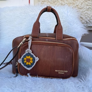 May include: A brown leather handbag with two top handles, a front zippered pocket, and gold-tone hardware. The text RE:DESIGNED PROJECT is printed on the front. A small, square, multi-colored crochet charm with a flower pattern hangs from the side.