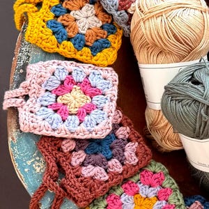 May include: A collection of square crochet motifs featuring multi-colored yarn patterns in shades of mustard yellow, pink, blue, and brown, arranged alongside two skeins of yarn in beige and sage green, showcasing intricate handmade stitch work.