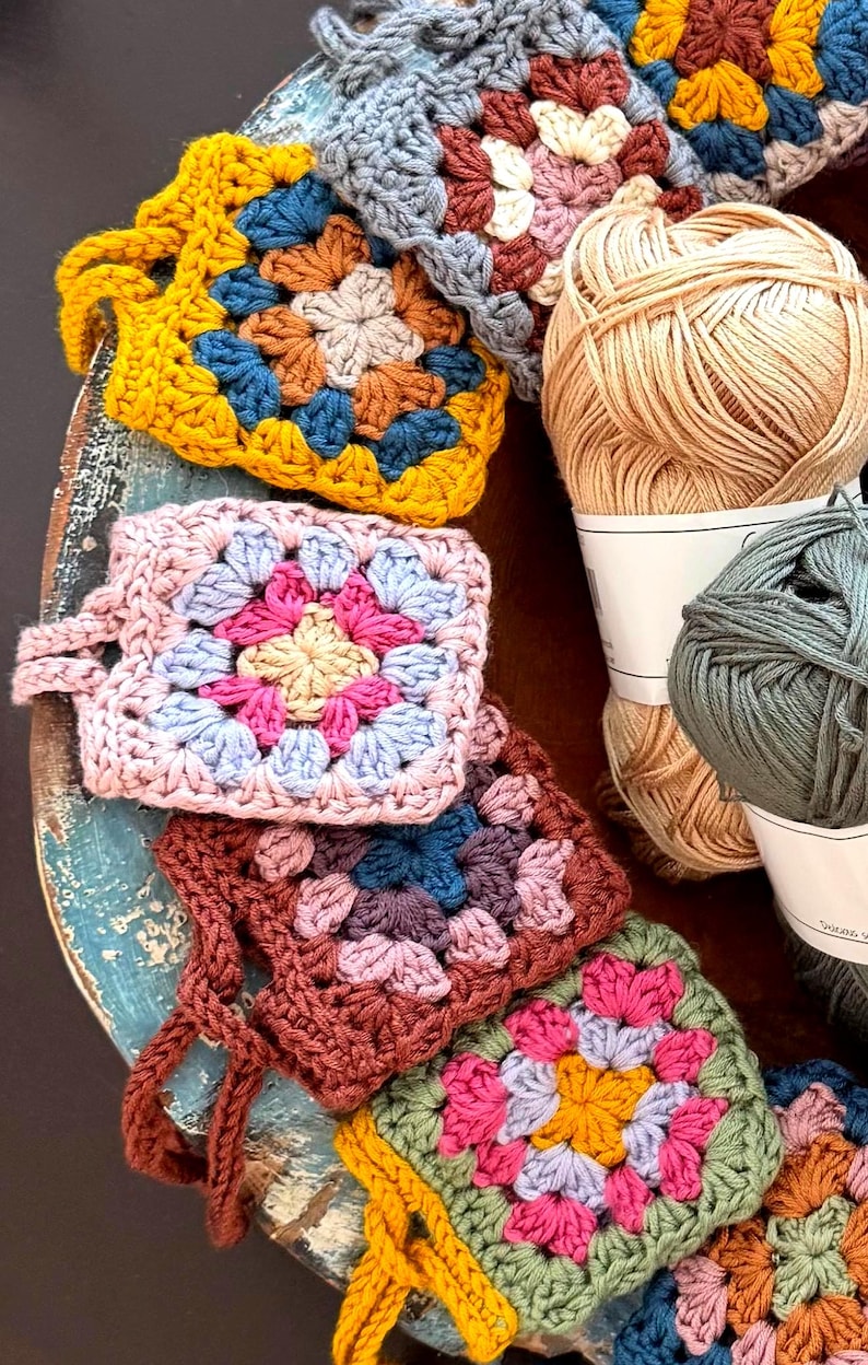 May include: A collection of square crochet motifs featuring multi-colored yarn patterns in shades of mustard yellow, pink, blue, and brown, arranged alongside two skeins of yarn in beige and sage green, showcasing intricate handmade stitch work.