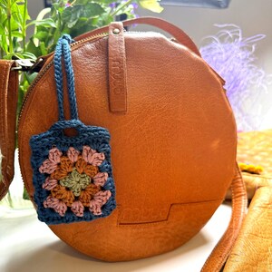 May include: A round, tan leather crossbody bag with a smooth texture and a zipper closure. Attached to the bag is a small, rectangular crochet charm featuring a granny square pattern in shades of blue, pink, green, and brown with a blue strap.