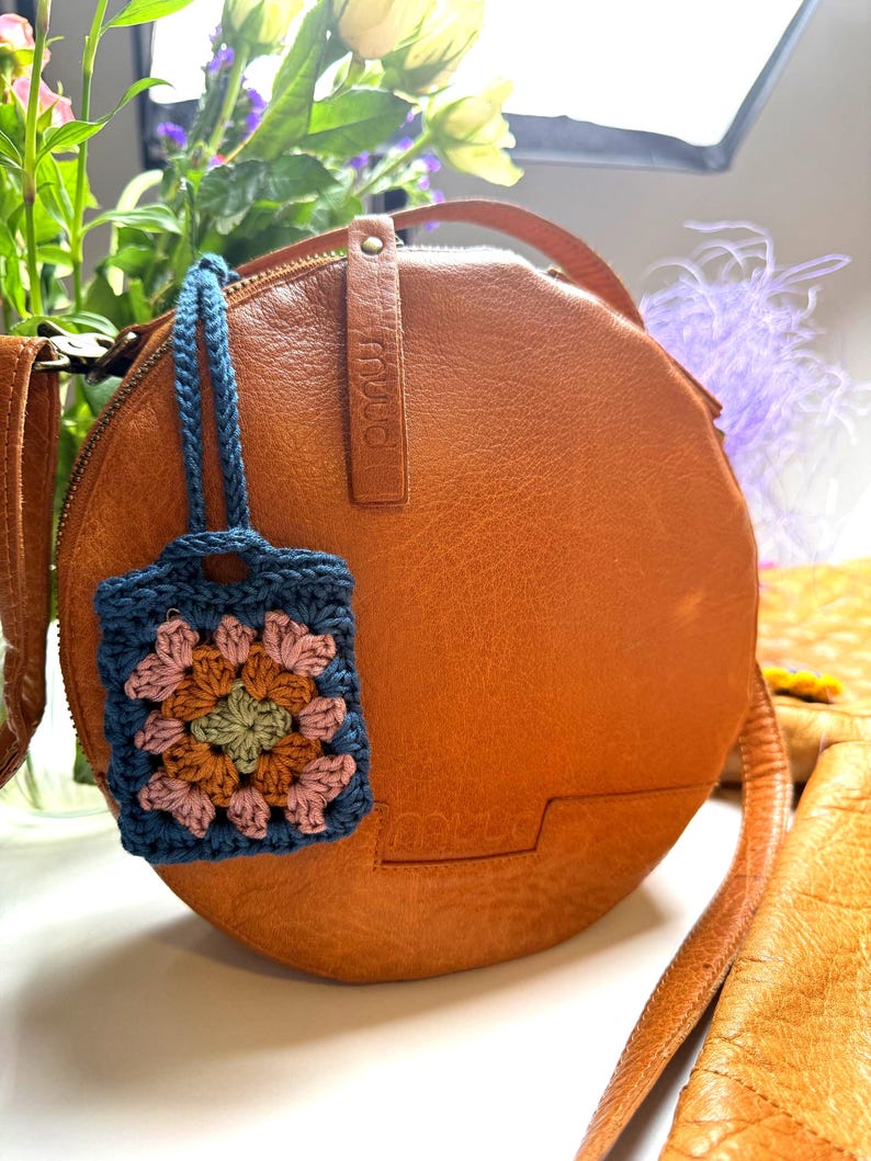 May include: A round, tan leather crossbody bag with a smooth texture and a zipper closure. Attached to the bag is a small, rectangular crochet charm featuring a granny square pattern in shades of blue, pink, green, and brown with a blue strap.