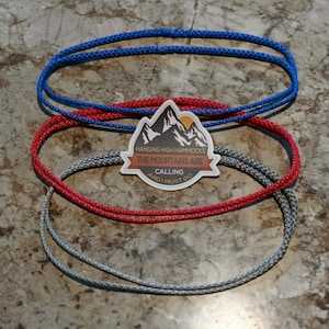 Puede incluir: Tres cuerdas de diferentes colores, gris, rojo y azul, están enrolladas en círculo sobre una superficie marrón texturizada. Una pegatina con el texto "Hanging High Hammocks The Mountains Are Calling And I Must Go" está en el centro de la imagen.