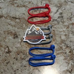 Könnte beinhalten: Drei Sätze Seil mit Knoten an den Enden. Die Seile sind rot, grau und blau. Ein Aufkleber mit einer Bergszene und dem Text "Hanging High Hammocks The Mountains Are Calling And I Must Go" befindet sich in der Mitte des Bildes.