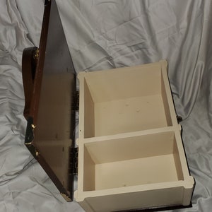 Può includere: Una scatola di legno con un coperchio incernierato e due scomparti. La scatola è marrone con un interno marrone chiaro. Il coperchio è aperto e la scatola è vuota.