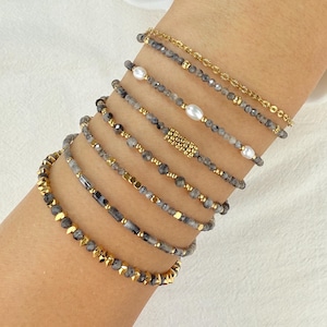 May include: A stack of seven bracelets featuring a mix of grey faceted beads, small white pearls, and gold-toned metallic accents. The set includes a thin gold chain bracelet, beaded strands with gold spacers, and a central gold-beaded cylinder.