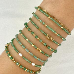 Può includere: Una collezione di braccialetti con perline verdi con dettagli dorati. I braccialetti presentano vari design, tra cui piccole perline, perline rettangolari e distanziatori dorati. Alcuni braccialetti includono anche dettagli di perle bianche.