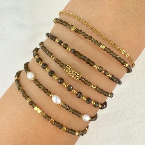 Puede incluir: Una colección de pulseras de cuentas doradas y verde oliva. Algunas pulseras presentan pequeñas cuentas doradas, mientras que otras tienen cuentas más grandes y facetadas. Una pulsera incluye perlas blancas. Las pulseras se muestran en una muñeca.