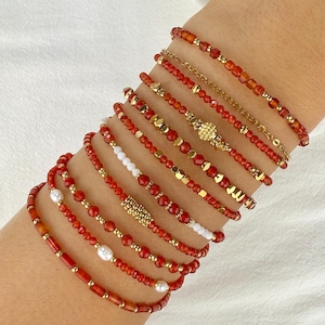 May include: A stack of ten bracelets featuring orange-red carnelian beads, small gold-tone metal beads, and white pearls. The bracelets vary in design, incorporating faceted stones, smooth cylindrical beads, and gold-tone spacers in a layered arrangement.