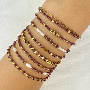 Peut inclure: Plusieurs bracelets de perles aux tons bordeaux et dorés. Certains bracelets comportent des perles blanches et des perles de culture. Les bracelets sont présentés sur un poignet, mettant en valeur les différents modèles et textures. Mesures en centimètres.