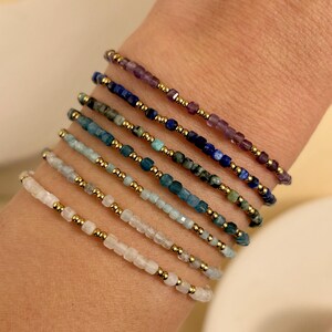 Peut inclure: Un empilement de sept bracelets de perles délicats composés de petites perles de pierres précieuses à facettes dans des tons de violet, bleu, sarcelle, bleu clair et blanc, alternées avec de minuscules perles métalliques dorées.