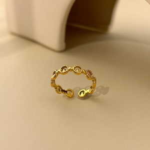 Pode incluir: Um anel ajustável dourado com um delicado design de elos de corrente. O anel é aberto na parte inferior e apresenta pequenos detalhes brilhantes. O anel é exibido em uma superfície de tons neutros.