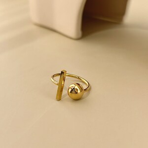 Puede incluir: Un anillo de oro con un diseño geométrico que presenta una esfera y una barra recta.