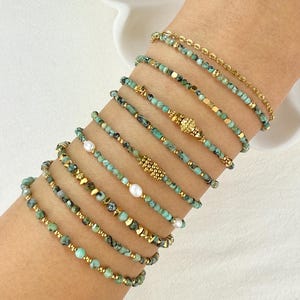 Pode incluir: Uma pilha de dez pulseiras com contas de pedra na cor turquesa, pequenas pérolas brancas e detalhes em metal dourado. As pulseiras variam em tamanho e textura, incorporando correntes douradas, espaçadores e pingentes decorativos de metal.