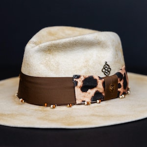 May include: A cream-colored fedora hat with a wide brim. The hat features a brown ribbon band with a leopard print accent and a decorative metal charm. Small beads are placed around the base of the hat.