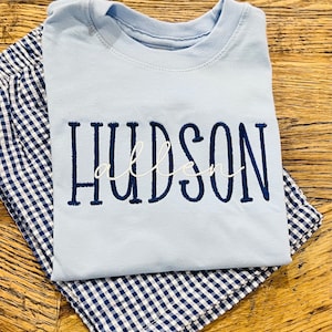 May include: A light blue t-shirt with the name "HUDSON" embroidered in navy blue, with "allen" embroidered in white script. The shirt is folded on top of navy and white gingham patterned fabric.