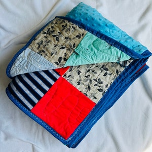 May include: A blue and red patchwork quilt with a variety of fabrics, including denim, striped fabric, and floral print. The quilt is folded in half and has a blue border.